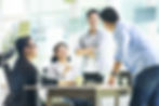 Four people in a bright office, smiling and discussing around a table. Light colors and greenery enhance the positive, collaborative mood.
