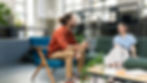 Man and woman chatting, seated in a modern office with plants. The man holds a cup, both are smiling. Bright, relaxed atmosphere.