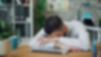 Man in checked shirt rests head on closed laptop, appearing tired. Background shows shelves and plants, creating an office setting.