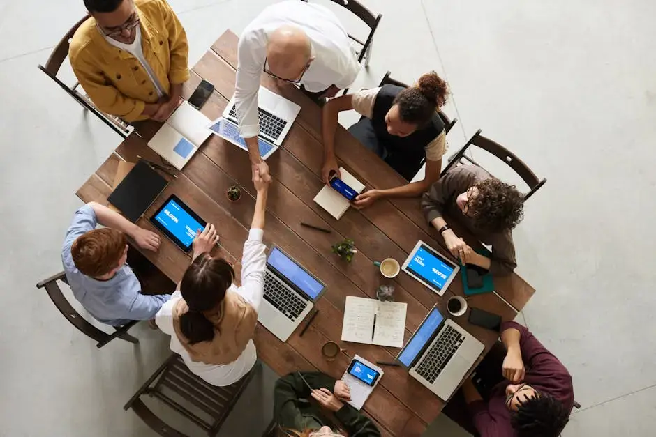 a group of 6 people sitting at a table with laptops and two people are shaking hands