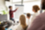 Man in a red sweater presents a flowchart on a flip chart to a group in an office. Attendees appear focused and engaged. Bright setting.