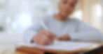 A person writes on paper with a pen, focusing intently. They're in a softly lit room, wearing a light sweater. Papers rest on a folder.